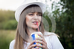 Portrait of young beautiful girl in hat with cocktail and straw on background of green summer nature