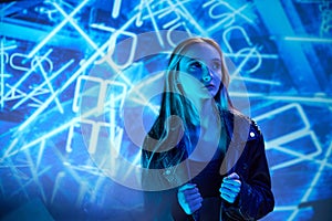 Portrait of young beautiful girl in leather jacket posing, looking away over blue background with abstract neon elements