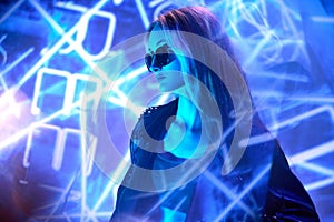 Portrait of young beautiful girl in leather jacket posing over blue background with abstract neon elements. Stylish