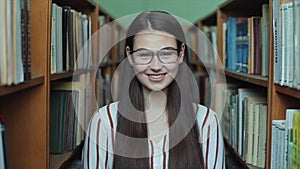 Portrait of young beautiful girl in library. Female student studying among lot of books between shelfs.
