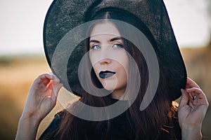 Portrait of a beautiful girl with pale skin and black lips in a big black hat. Woman in the image of a witch for Halloween.