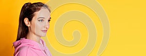 Portrait of young beautiful girl posing in pink hoodie over yellow studio background. Banner, flyer. Concept of youth