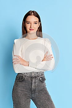 Portrait of young beautiful girl, student in white blouse and jeans posing over blue studio background. Concept of