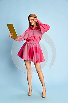 Portrait of young beautiful girl in stylish pink dress and glasses posing isolated over blue background. Business woman