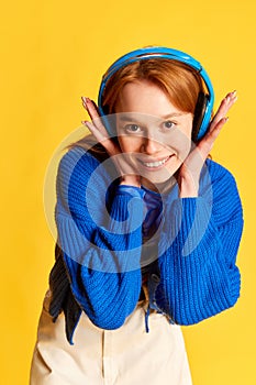 Portrait of young beautiful girl, teenager with red hair wearing headphones and smiling over yellow studio background