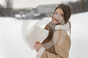 Portrait of a young beautiful girl in a white coat holding a hat in hands on a background of winter park