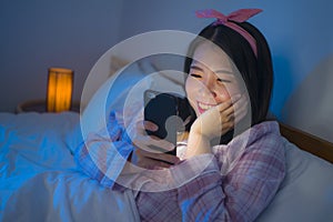 Portrait of young beautiful and happy sweet Asian Chinese girl with bow headband and pajamas networking with mobile phone in bed