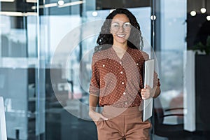 Portrait of young beautiful hispanic female student with laptop, woman at work in modern university classroom, smiling