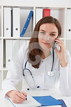 Portrait of a young beautiful medical doctor talking on the phone and writing something in a notebook.