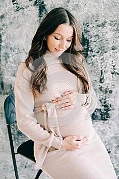 Portrait of a young beautiful pregnant woman in a beige dress sitting on a chair near a gray wall and holding her stomach