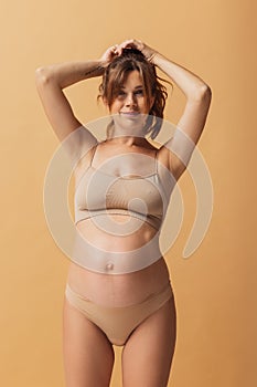 Portrait of young beautiful pregnant woman in lingerie isolated over sand color studio background. Natural beauty, happy