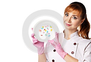 Portrait of young beautiful red-haired woman doctor or nurse in white special uniform and gloves showing plate with colorful pills