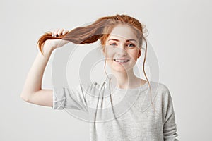 Portrait of young beautiful redhead girl touching her hair tail looking at camera over white background.