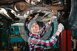 Portrait of a young beautiful smiling female mechanic in uniform and gloves who repairs a car. The car is on the lift