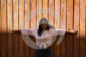 Portrait of young and beautiful South American woman in white shirt, jeans and sunglasses with open arms on a wooden wall, half