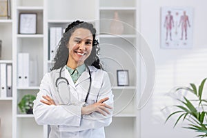 Portrait of young beautiful successful female doctor inside medical office, hispanic woman with curly hair in white