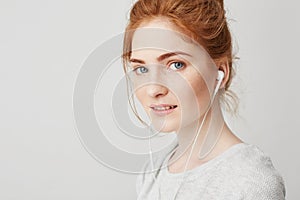 Portrait of young beautiful tender redhead girl with blue eyes in headphones looking at camera smiling over white