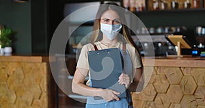 Portrait of young beautiful woman barista in medical mask standing near the bar and holding menu