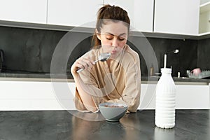 Portrait of young beautiful woman in bathrobe, eating cereals for breakfast, leans on kitchen worktop, looking at her
