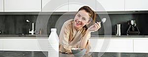 Portrait of young beautiful woman in bathrobe, eating cereals for breakfast, leans on kitchen worktop, looking at her