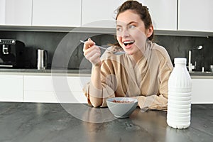 Portrait of young beautiful woman in bathrobe, eating cereals for breakfast, leans on kitchen worktop, looking at her