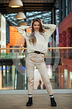 Portrait of a young beautiful woman in beige sweater and pants posing in the mall