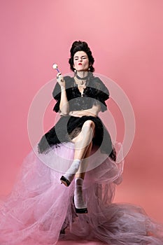 Portrait of young beautiful woman in black evening little dress sitting on fabric cloud isolated on pink background