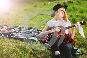 Portrait of a young beautiful woman in a black hat. Girl sitting on the grass and playing guitar