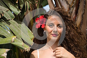 Portrait of a young and beautiful woman, blonde, with curly hair and blue eyes, next to some palm trees, with a red Pacific flower