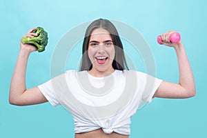 Portrait of young beautiful woman with broccoli and dumbbell, over blue background. Healthy food, health life.