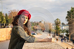 Portrait of a young and beautiful woman, brunette, with curly hair, with a green jacket and red beret, leaning on a stone wall.