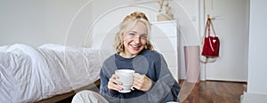 Portrait of young beautiful woman in casual clothes, sitting on bedroom floor with cup of tea, drinking and smiling