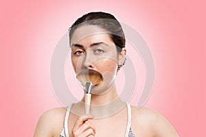 Portrait of a young beautiful woman, gravely holding a fluffy brush in the form of a mustache. Pink background. The concept of photo