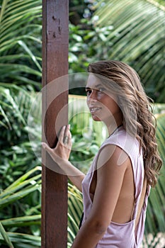 Portrait of young beautiful woman in pink dress with open back on balcony of tropical bungalow with palm trees view