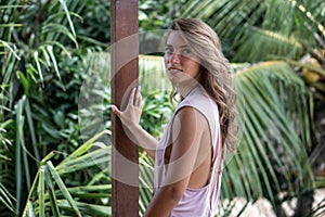 Portrait of young beautiful woman in pink dress with open back on balcony of tropical bungalow with palm trees view