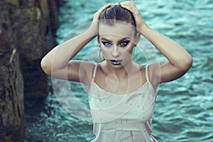 Portrait of young beautiful woman with provocative make up standing in the sea water under the rain and touching her wet hair