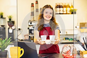 Portrait of a young beautiful woman in red apron, keeps white cup in hand, smiling, looking at camera, on a coffee shop