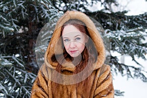 Portrait of young beautiful woman with red hair and blue eyes in faux fur coat on background of winter snowy park