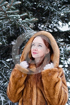 Portrait of young beautiful woman with red hair and blue eyes in faux fur coat on background of winter snowy park