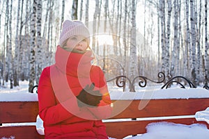 Young beautiful woman in red warm jacket sits on bench in pictorial park with birches and warms hands in winter frosty sunny day