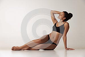 Portrait of young beautiful woman sitting on floor, posing in black underwear  over gray background. Perfect