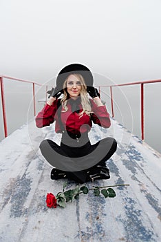 Portrait of young beautiful woman sitting on the pier and holding red rose in winter in fog