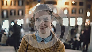 Portrait of young beautiful woman standing in the city centre in evening. Student girl looks at camera, smiling.