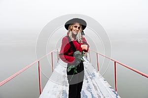 Portrait of young beautiful woman standing on the pier and holding red rose in winter in fog