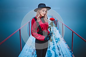 Portrait of young beautiful woman standing on the pier and holding red rose in winter in fog