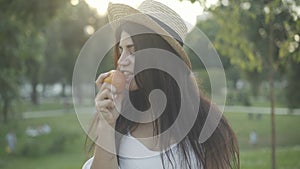 Portrait of young beautiful woman in straw hat eating healthful apple in sunny park and smiling at camera. Attractive