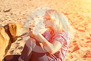 Portrait of young beautiful woman in sunglasses sitting on sand beach hugging golden retriever dog. Girl with dog by sea