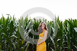Portrait of young beautiful woman wearing a yellow dress standing in a green corn field. Summertime and lifestyle