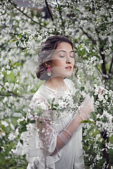 portrait of a young beautiful woman in white clothes standing next to a blooming cherry tree in spring.
