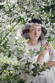 portrait of a young beautiful woman in white clothes standing next to a blooming cherry tree in spring. a wreath on the
